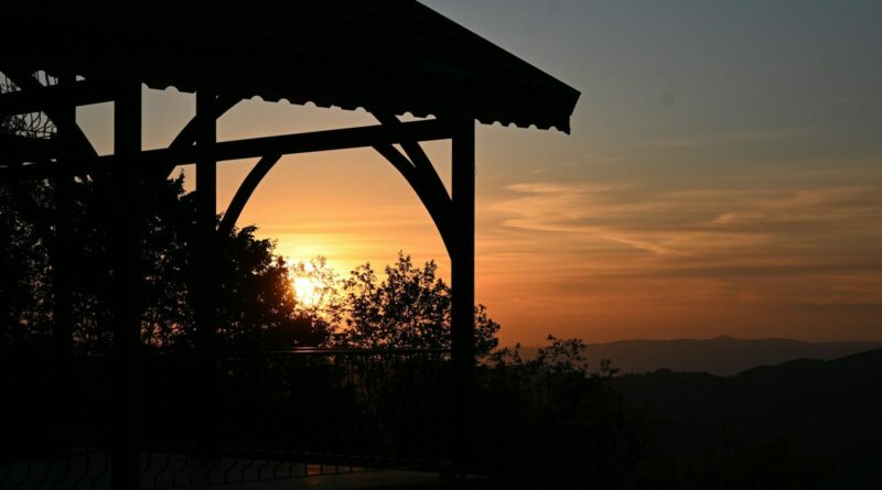 A wooden gazebo silhouette against a warm orange sky and distant mountain ridges